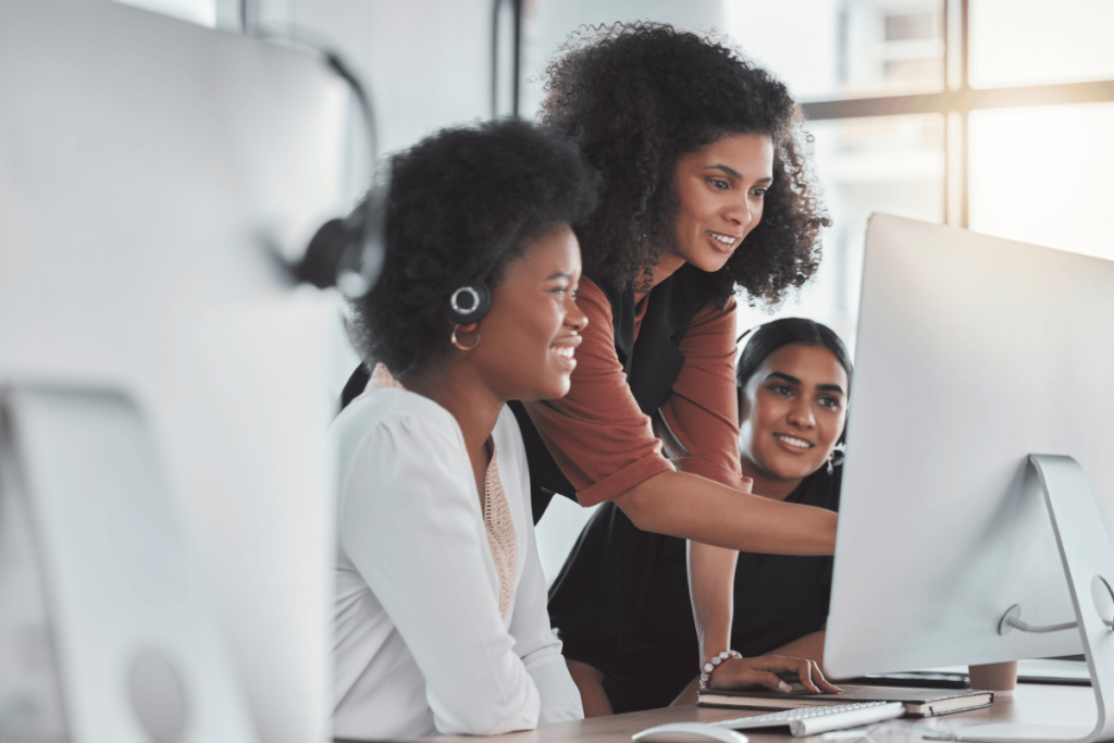 4 Great Coaching Options You Can Use to Grow Your Contact Center. A female coach helps two agents at a call center and points to a computer screen.