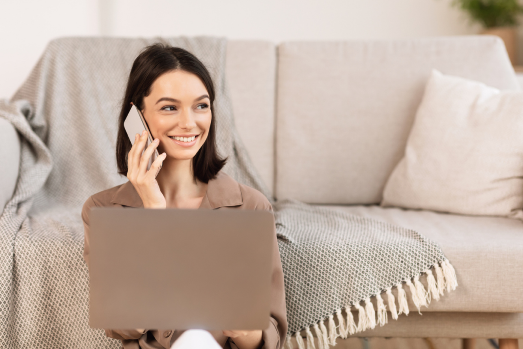 What Does “The Customer is Always Right” Really Mean? A smiliing young woman sits on the floor in front of a couch on her computer and holds a phone to her ear.