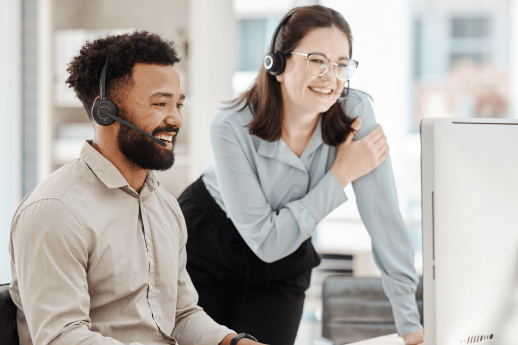 A professional woman wearing a headset coaches a man sitting at a computer and wearing a headset at a call center