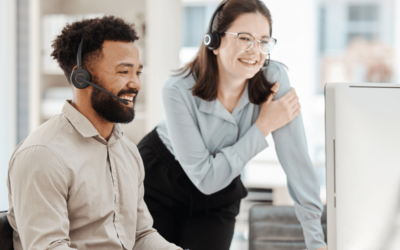 A professional woman wearing a headset coaches a man sitting at a computer and wearing a headset at a call center