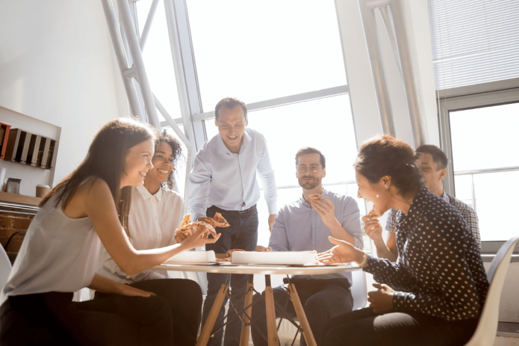 happy co-ed group of professional coworkers smile as they sit around the work table and eat pizza in a well lit room with windows