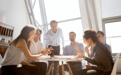 happy co-ed group of professional coworkers smile as they sit around the work table and eat pizza in a well lit room with windows