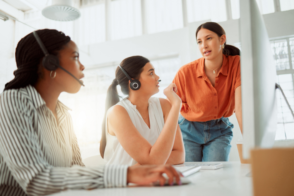 How to Get Started with Quality Assurance Coaching and Training Programs. Two women at a call center wear headphones and listen to a third woman coaching them.